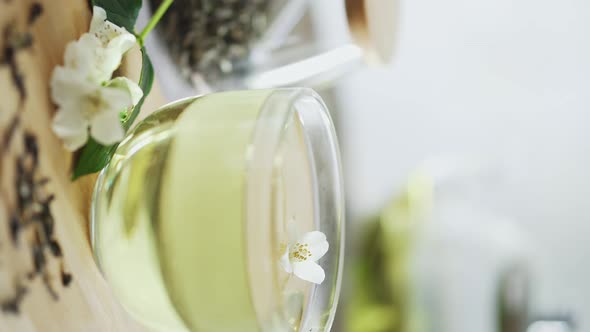 Glass Cup Of Green Tea With Jasmine On Bamboo Table With Sprig Of Jasmine. Hand Puts Fresh Jasmine alt