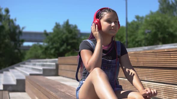 The Girl Listens to the Music on the Headphones Saddle on the Bench in the Park alt