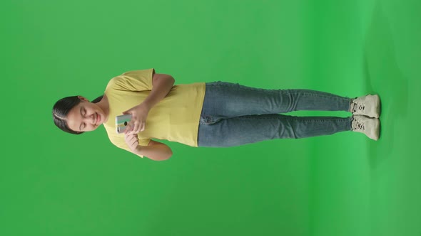 Full Body Of Smiling Kid Girl Use Mobile Phone And Pointing To Smart Phone In Green Screen Studio alt