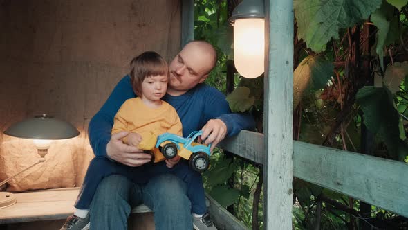 Father is Playing on Porch of the House with His Cute Little Child in a Toy Car alt