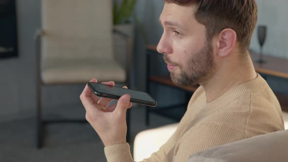 Man sitting on sofa holds phone talks on speakerphone, audio message alt