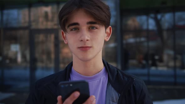 Medium Shot of Handsome Young Man Working on Smartphone Stands on the Street in Front of a Modern