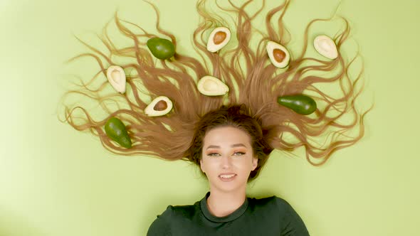 top view woman lying on colored background with avocado fruits on long hair, cosmetics, beauty alt