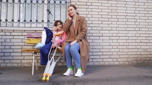 Mom Talking On Phone Daughter With Crutches On Bench In Yard Stock Footage