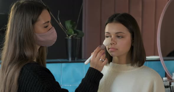 Female MakeUp Artist in Protection Mask Using Brush to Work with the Face of Client Young Woman on