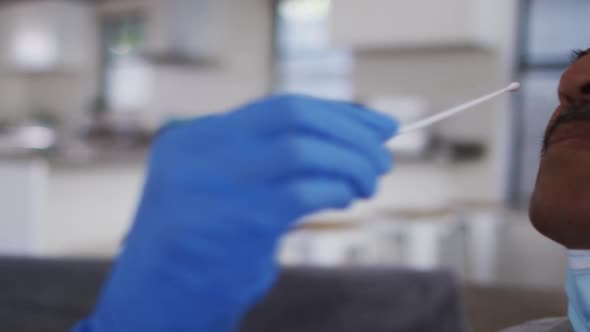 Mixed race female doctor wearing mask doing swab test on senior man at home alt