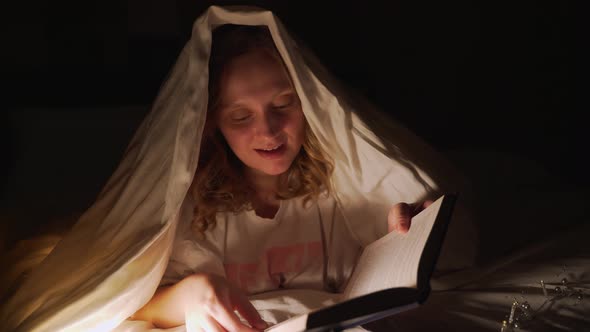 Woman Reads Book Under Blanket in the Dark and Illuminates the Pages of the Book with Flashlight