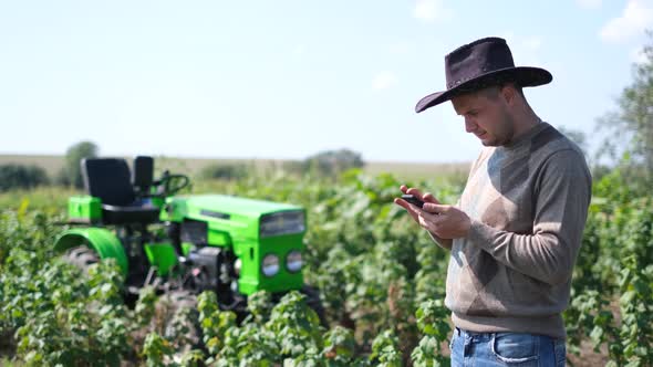 A Young Agricultural Worker Holds a Smartphone While He is Working Near His Tractor alt