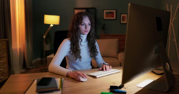 A Beautiful Young Girl with Curly Hair Sits in Front of Computer Screen in Evening alt