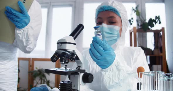 Happy Female Clinic Lab Scientist in Protection Suit and Shield Holding Flask with New Vaccine alt