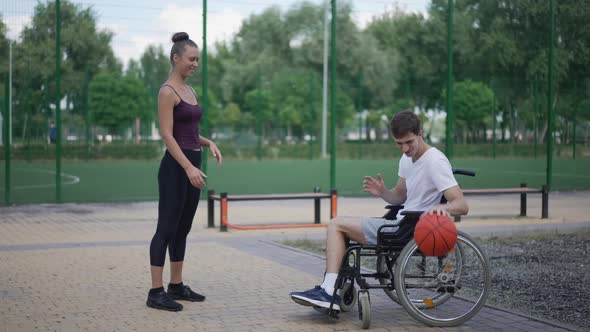 Wide Shot Happy Girlfriend Playing Basketball with Boyfriend in Wheelchair Outdoors alt