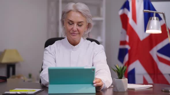 Positive Caucasian Middle Aged Woman Sitting at Table with Tablet Waving Greeting Talking Smiling alt