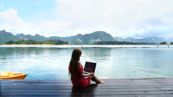 Woman Freelancer Sitting on Wooden Pier on Lake and Work on Project on Laptop alt