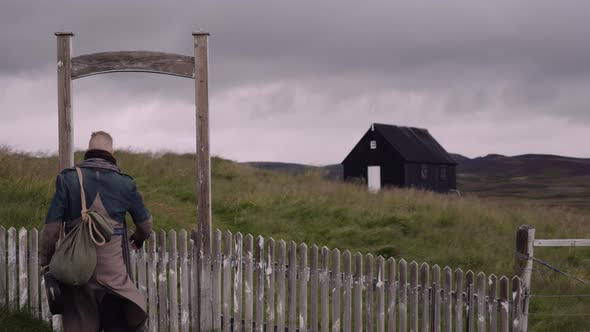 Stylish Man with a Sack Bag and Banjo Case Walks in Through Wooden Fence Gate alt