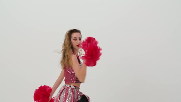 Young Cheerleader with Red Pompoms in Uniform is Dancing on White Background in Studio alt