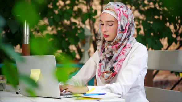 Muslim Student in Hijab Writes an Essay on a Laptop in the Park on a Summer Day alt