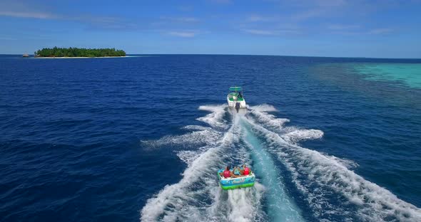 Aerial drone view of man and woman on an inflatable tube towing behind a boat to a tropical island alt