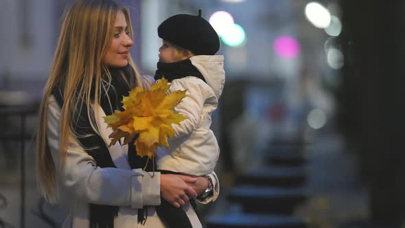 Mom with her little daughter in her arms walks on the evening street in the city in autumn