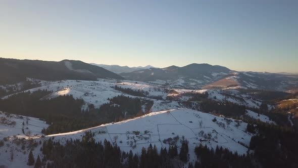 Aerial view of small village with scattered houses on snow covered hills in winter and bare dark alt