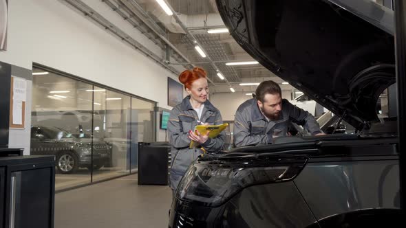 Two Professional Car Mechanics Examining Engine of a Car, Making Notes alt