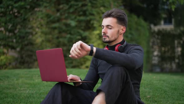 Young Male Freelancer Sitting on Park Meadow Surfing Internet on Laptop Checking Time on Watch alt