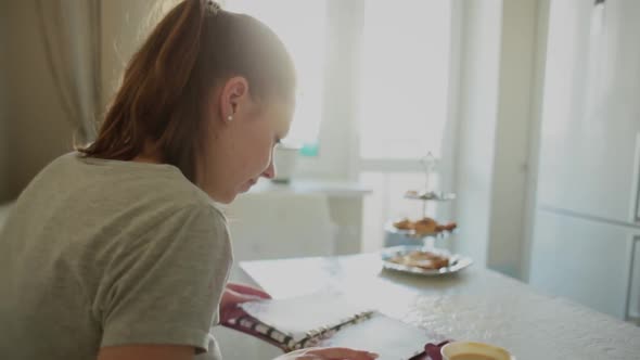 Girl Kitchen Opposite the Window at the Table Leafs Through a Notebook