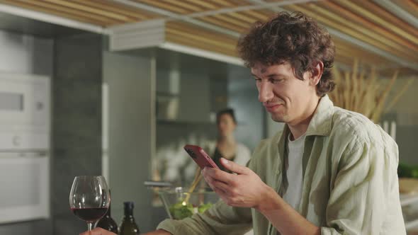 Man Drinking Wine and Using Mobile While Woman Cooking alt