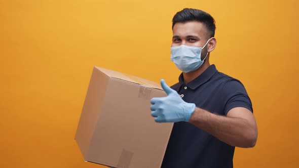 Indian Man with Face Mask and Gloves Holding Cardboard Box and Showing Thumb Up alt