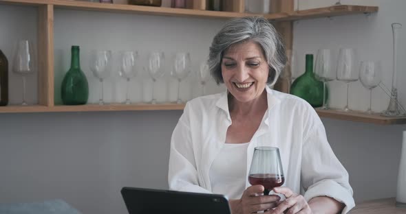 Mature Elderly 60 Year Old Woman Standing with a Tablet Drinking Wine and Enjoying Video Call