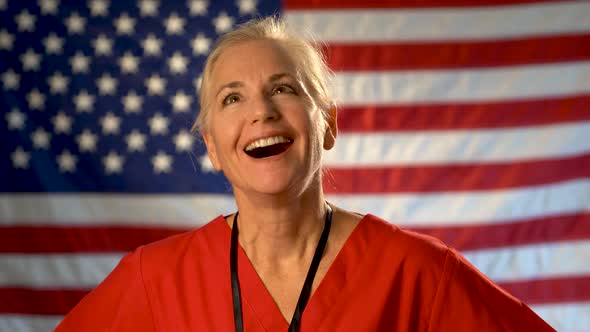 Medium tight portrait of nurse looking at the camera, relieved and very happy with a US flag behind alt