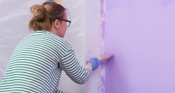 The Painter Girl Paints the Wall with a Roller and a Brush in Lilac Color alt