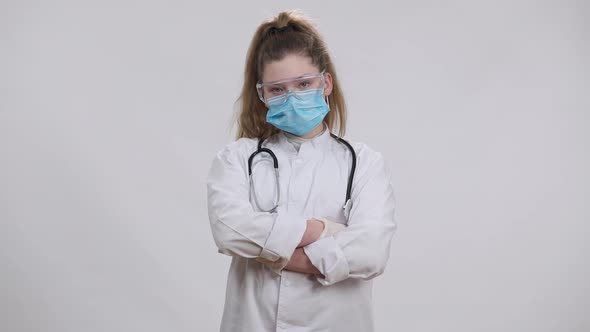 Little Nurse Doctor in Coronavirus Face Mask Medical Gown and Eyeglasses Crossing Hands Posing at alt