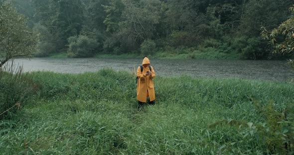 Hiker in a Yellow Raincoat with a Backpack in the Tall Grass Next to the River
