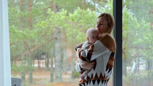 mother hugs baby by the window overlooking a pine forest