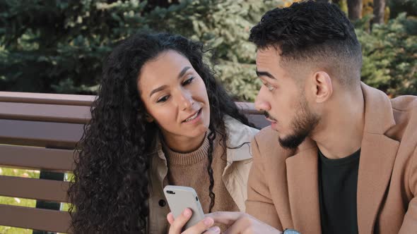 Hispanic Couple Sit on Bench in Autumn Park Young Guy Hold Phone Man and Girl Talk Look at Screen alt