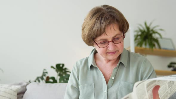 A Closeup View of a Beautiful Mature Woman is Reading a Newspaper Sitting on Sofa at Home in the alt