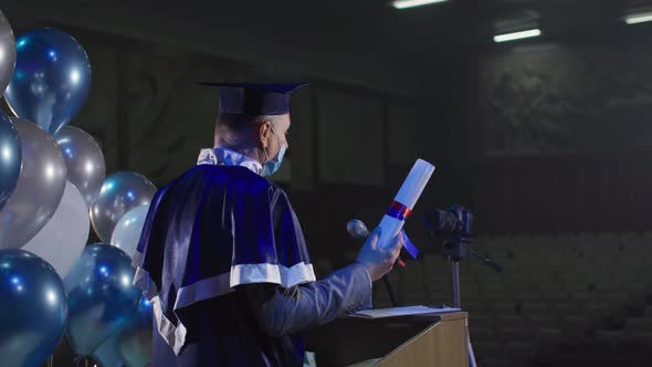 University Lecturer in Academic Clothes and Hat in Medical Mask is Presenting Diplomas Using Video alt