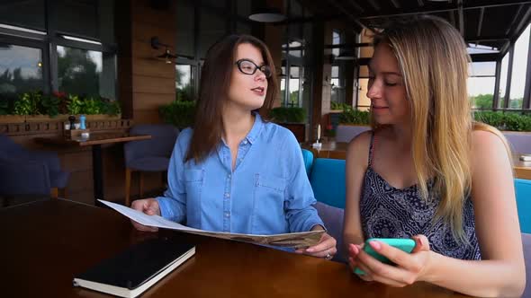 Female Friends Sitting at Restaurant and Looking at Menu and Smartphone Screen.