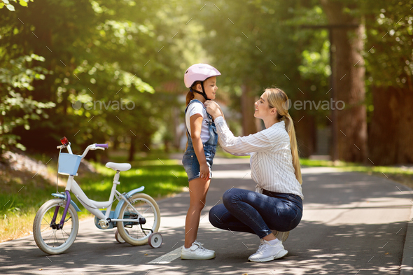 Mother Putting Helmet On Daughter Teaching To Ride Bicycle Outdoor