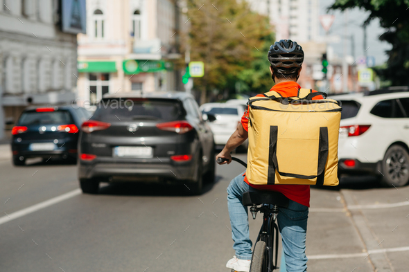 Male courier with helmet and big yellow backpack ride on bicycle along road on street - Stock Photo - Images