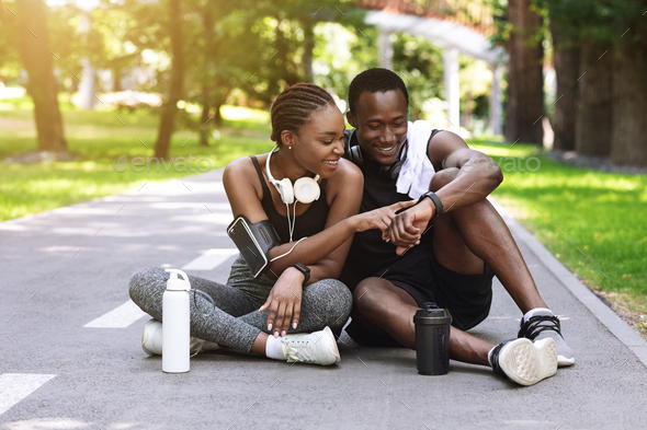 Joyful black couple resting after jogging in park, checking smartwatch