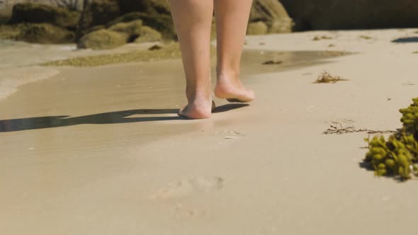 A close up shot of the heels of a young female walking leisurely across a beach, shallow ocean waves alt