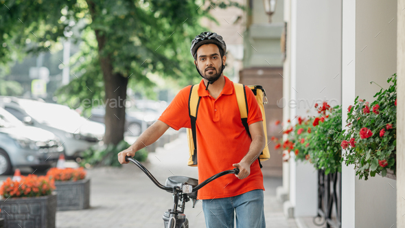 Bike, city and food delivery concept. Courier with beard, in helmet and bag, walks with bicycle down