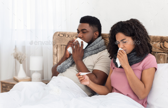 Sick black couple sitting in bed, measuring fever, sneezing noses