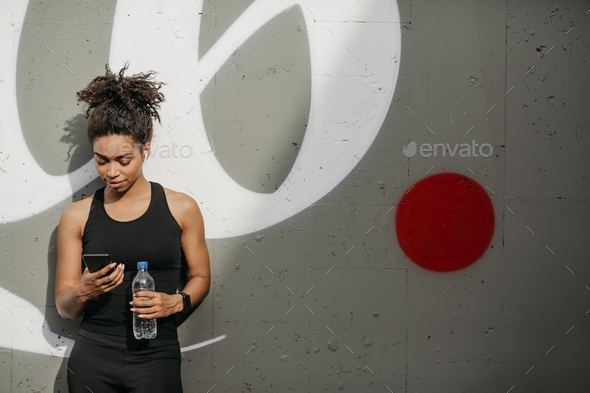 African american woman in sportswear with headphones, fitness tracker and bottle looks at smartphone