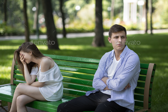 Frustrated young guy and crying girl after fight sitting on bench at park