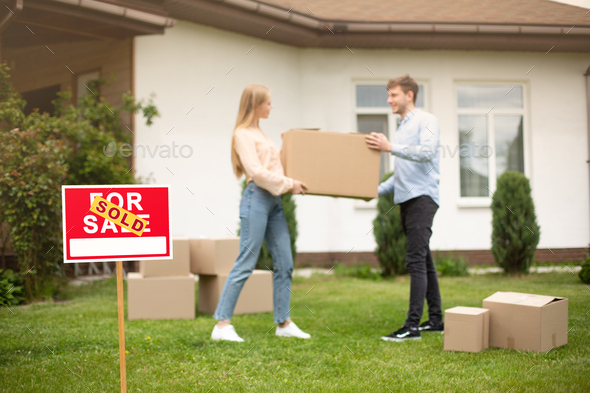 Millennial couple with carrying box in front of new home on moving day, focus on SOLD sign. Copy