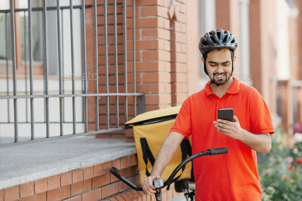 Delivery address. Smiling courier in uniform and helmet with backpack and bike watching at - Stock Photo - Images