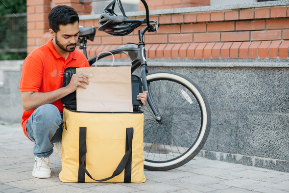 Courier and package for the client. Busy guy courier in uniform with bicycle puts goods in bag