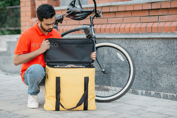 Packing the order in thermal bag. Serious guy courier with beard in uniform with bicycle looks at